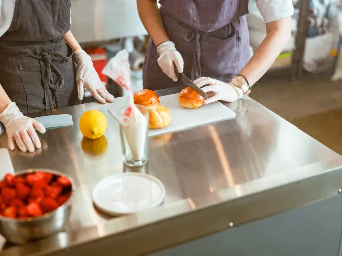 Dos personas con delantales y guantes preparando bollos en una cocina profesional, cortando pan sobre una mesa de acero inoxidable.