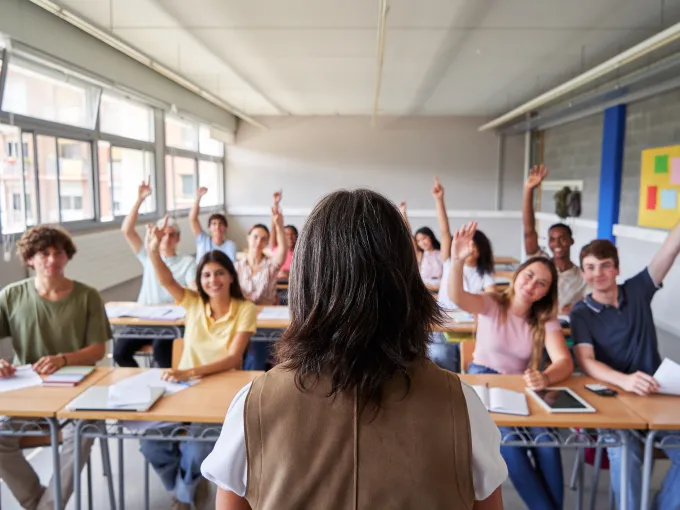 Estudiantes de secundaria en un aula levantando la mano mientras la docente los observa desde el frente.