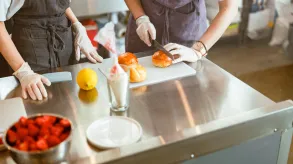 Dos personas con delantales y guantes preparando bollos en una cocina profesional, cortando pan sobre una mesa de acero inoxidable.