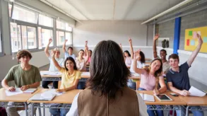 Estudiantes de secundaria en un aula levantando la mano mientras la docente los observa desde el frente.