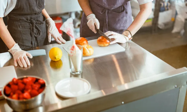 Dos personas con delantales y guantes preparando bollos en una cocina profesional, cortando pan sobre una mesa de acero inoxidable.
