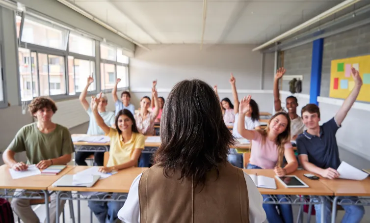 Estudiantes de secundaria en un aula levantando la mano mientras la docente los observa desde el frente.