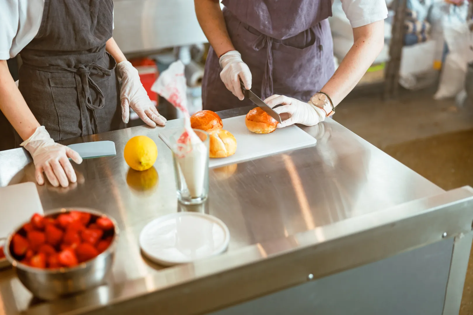 Dos personas con delantales y guantes preparando bollos en una cocina profesional, cortando pan sobre una mesa de acero inoxidable.